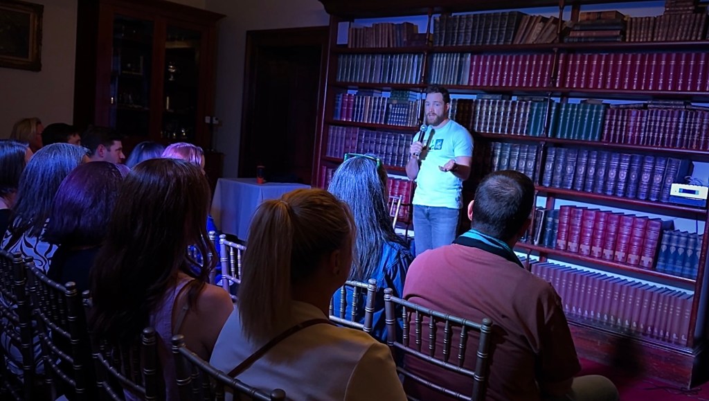 Russell is standing in front of a large bookshelf filled with old stately books. He is holding out his open hand in front of him, like he is handing something over. There is an audience in front of him, their faces are hidden but I can guarantee they are having a good time. 